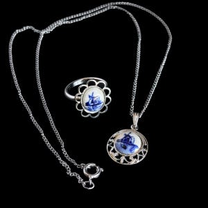 May include: A silver necklace and ring set with a Delftware design. The ring has a scalloped edge and the pendant is round. Both feature a blue and white windmill scene. The jewellery is displayed on a black background.