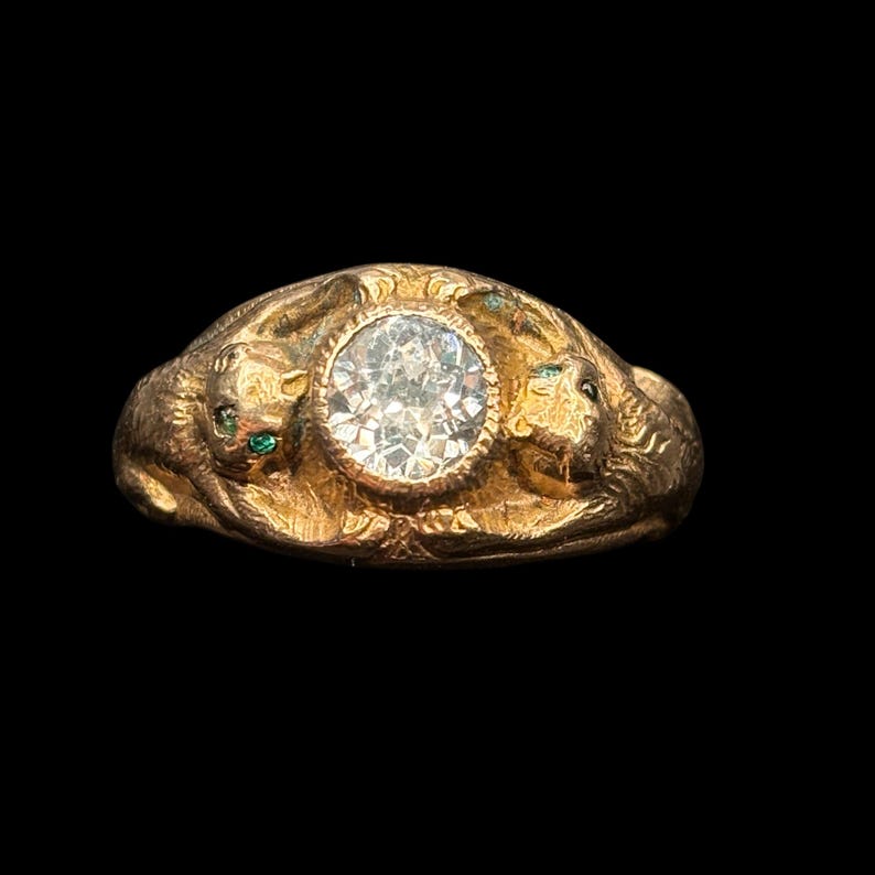 May include: A gold ring with a large, clear, round-cut diamond at the center. The ring features detailed relief carvings of two lion-like creatures on either side of the diamond, with small green accents. The ring is set against a black background.