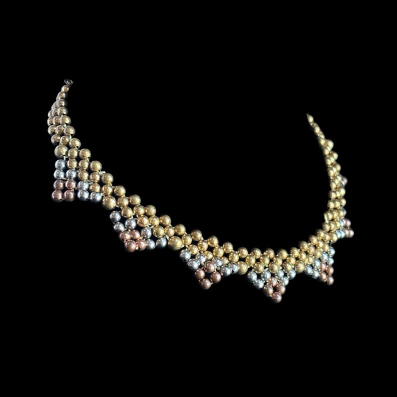 May include: A necklace featuring gold, silver, and rose gold beads. The necklace is made up of rows of small, round beads in a zig-zag design. The necklace is set against a black backdrop.