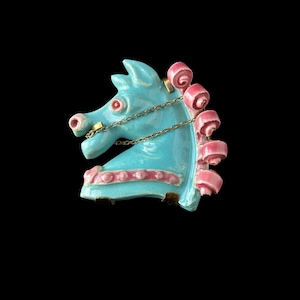 May include: A turquoise horse head brooch with pink accents and a silver chain bridle. The brooch has a glossy finish and a vintage aesthetic. The horse's mane is adorned with pink, curled details, and the eyes are red.
