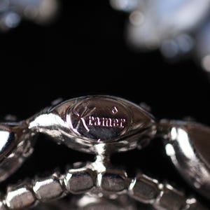 May include: Close-up of a silver metal bracelet with a clear rhinestone design. The bracelet has a small, oval-shaped tag with the word "Kramer" engraved on it.