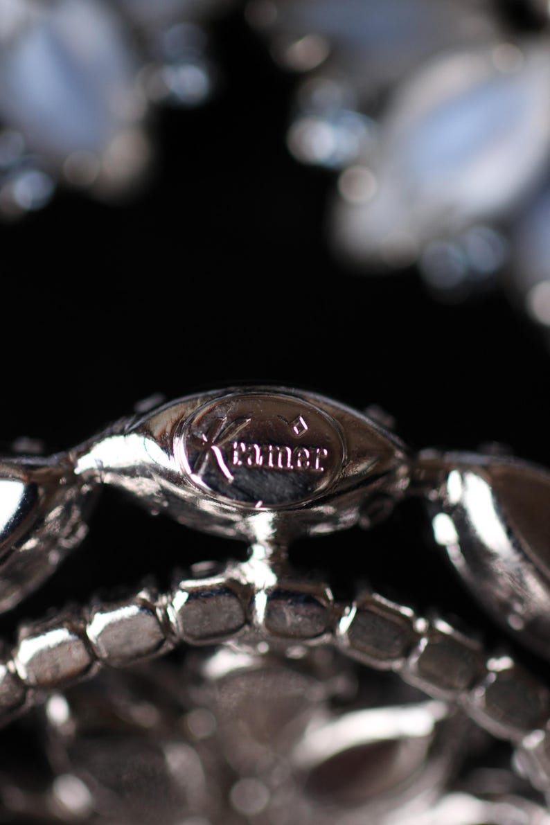 May include: Close-up of a silver metal bracelet with a clear rhinestone design. The bracelet has a small, oval-shaped tag with the word "Kramer" engraved on it.