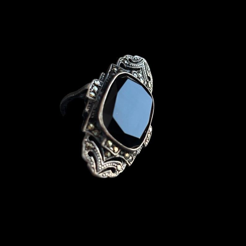 K&ouml;nnte beinhalten: Ein Ring im Vintage-Stil mit einem gro&szlig;en, facettierten schwarzen Edelstein. Der Ring hat eine verzierte, silberfarbene Fassung mit aufwendigem Filigran und kleinen, funkelnden Akzenten. Der Edelstein ist quadratisch und vor schwarzem Hintergrund platziert.