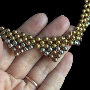 May include: A multi-strand necklace with small, round beads in gold, silver, and copper hues. The necklace is held in a hand against a black backdrop, highlighting the design and colour variations of the jewellery.