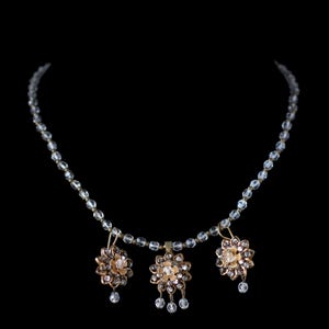 Peut inclure: Un collier délicat avec trois pendentifs floraux. Les pendentifs sont dorés avec des accents de cristaux transparents et des gouttes de cristal pendantes. La chaîne du collier est faite de petites perles transparentes. Le fond est noir.