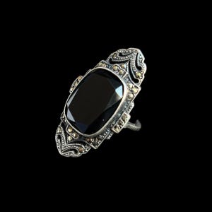 K&ouml;nnte beinhalten: Ein silberfarbener Ring mit einem gro&szlig;en, rechteckigen schwarzen Edelstein. Der Ring weist aufwendige Details und kleine, runde Akzentsteine auf. Das Design ist vor einem schwarzen Hintergrund platziert, der die komplizierten Details des Rings hervorhebt.