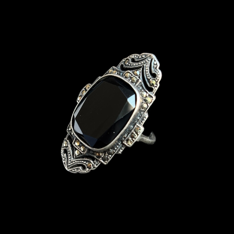 K&ouml;nnte beinhalten: Ein silberfarbener Ring mit einem gro&szlig;en, rechteckigen schwarzen Edelstein. Der Ring weist aufwendige Details und kleine, runde Akzentsteine auf. Das Design ist vor einem schwarzen Hintergrund platziert, der die komplizierten Details des Rings hervorhebt.