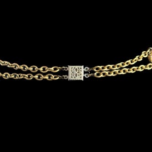 May include: A gold-coloured necklace featuring two parallel chains. The chains are joined by a square, silver-coloured clasp with a floral pattern. The background is black.