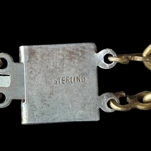 May include: Close-up of a silver clasp with the word "STERLING" engraved. The clasp is rectangular and connects to a gold chain. The background is black.