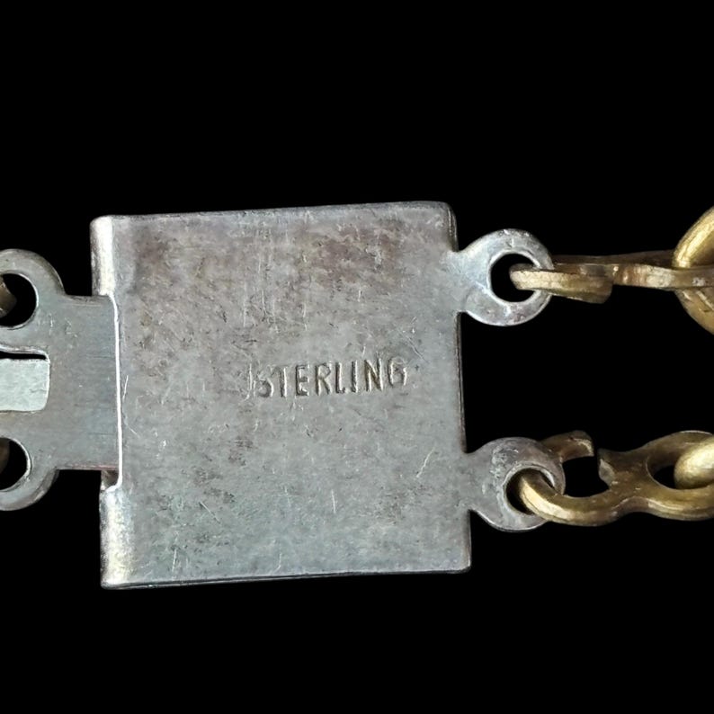 May include: Close-up of a silver clasp with the word "STERLING" engraved. The clasp is rectangular and connects to a gold chain. The background is black.