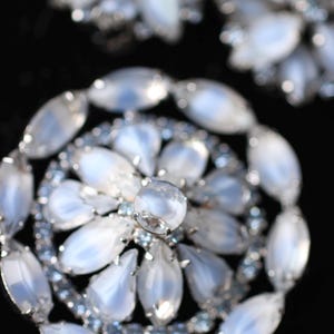 May include: A large, silver-toned brooch with a circular design. The brooch is made of many oval-shaped, iridescent white stones set in silver. The center of the brooch features a larger, round, iridescent white stone.