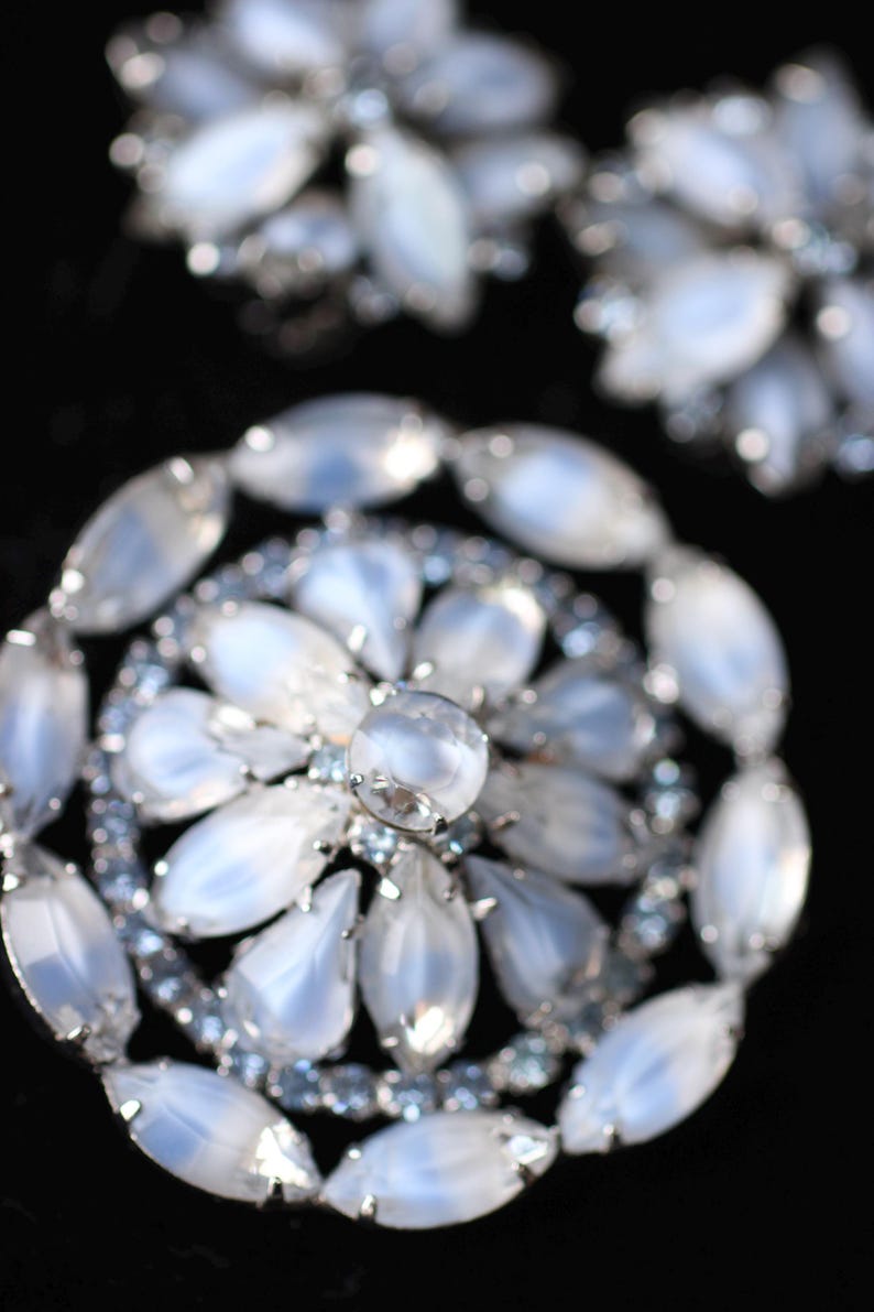 May include: A large, silver-toned brooch with a circular design. The brooch is made of many oval-shaped, iridescent white stones set in silver. The center of the brooch features a larger, round, iridescent white stone.