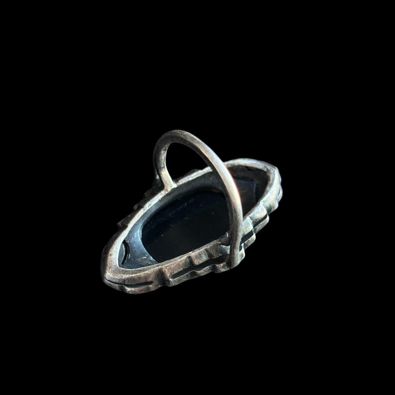 K&ouml;nnte beinhalten: Ein silberfarbener Ring mit einem ovalen, schwarzen Stein. Der Ring hat ein dekoratives, strukturiertes Band und ein einfaches, gew&ouml;lbtes Band. Der Ring ist vor einem schwarzen Hintergrund platziert.