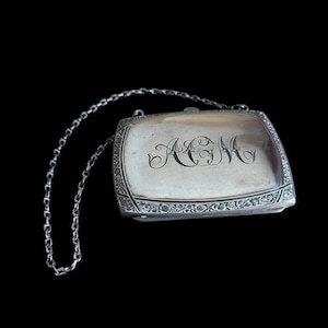 May include: A silver rectangular purse with a chain handle. The purse has ornate detailing around the edges and features the engraved initials "A&M" on the front. The background is solid black.