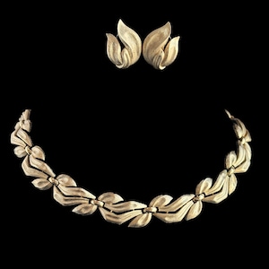 Puede incluir: Un conjunto de joyas en tono dorado con un collar y pendientes a juego. El collar tiene un diseño en forma de hoja, y los pendientes tienen forma de flores estilizadas. El conjunto se muestra sobre un fondo negro.