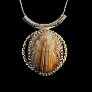 May include: A gold-toned necklace with a pendant featuring a brown and cream-colored oval stone. The stone is carved with a detailed design and is surrounded by a gold-colored twisted rope-like border. The necklace has a curved bar at the top.