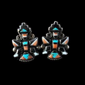 May include: A pair of silver-toned stud earrings with a geometric design. The earrings feature inlaid turquoise, black, white, and orange stones. The design has a Southwestern or Native American aesthetic.
