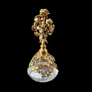 May include: A clear glass perfume bottle with a faceted base and a gold-toned metal overlay featuring intricate rose and leaf designs. The stopper is a tall, ornate gold-toned metal sculpture of a rose bouquet with multiple leaves and stems.