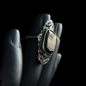 K&ouml;nnte beinhalten: Ein silberfarbener Ring mit einem l&auml;nglichen, rechteckigen schwarzen Edelstein. Der Ring hat aufwendige Filigranarbeiten und kleine, funkelnde Akzente. Der Ring wird vor schwarzem Hintergrund pr&auml;sentiert.