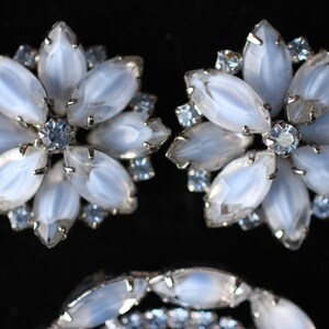 May include: A pair of silver tone earrings with a flower design. Each earring has a cluster of white iridescent stones surrounded by smaller clear stones.