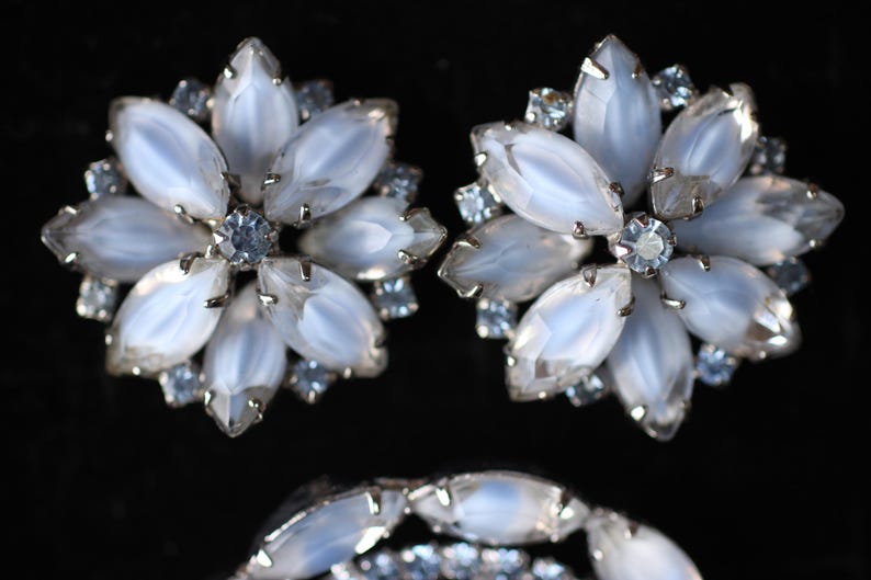 May include: A pair of silver tone earrings with a flower design. Each earring has a cluster of white iridescent stones surrounded by smaller clear stones.