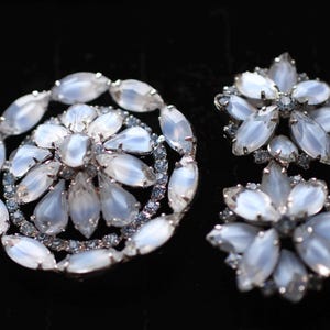 May include: A set of two silver tone brooches with white iridescent glass stones. The larger brooch is round and features a flower design. The smaller brooch is a starburst shape.