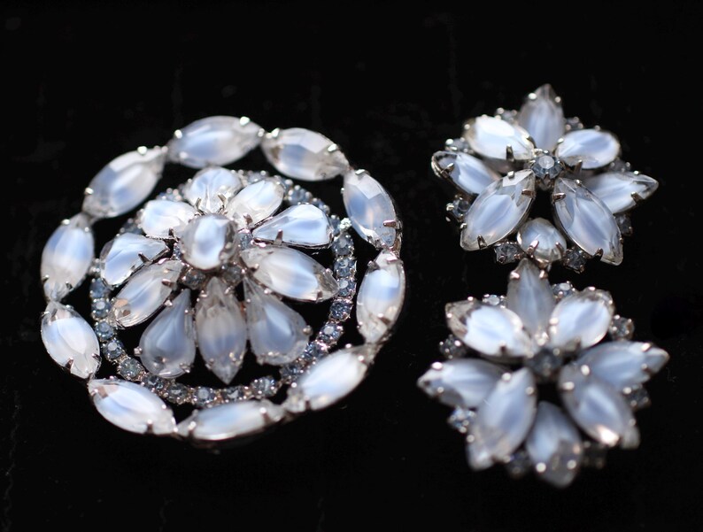 May include: A set of two silver tone brooches with white iridescent glass stones. The larger brooch is round and features a flower design. The smaller brooch is a starburst shape.