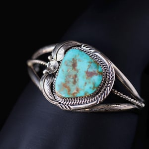 May include: Silver cuff bracelet featuring a large, oval turquoise stone with brown matrix. The stone is set in a decorative bezel with a braided edge. The bracelet has leaf and floral accents.