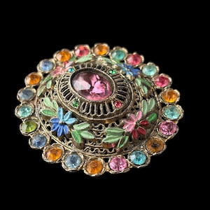 Op de afbeelding: Vintage broche met een roze ovale edelsteen in het midden, omringd door een filigraan ontwerp en kleurrijke edelstenen. De broche heeft geschilderde bloemaccenten in blauw, roze en groen, op een metalen basis. Een decoratief sieraad.