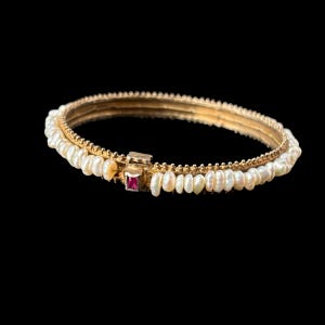 May include: A gold bangle bracelet featuring a row of small white pearls along the outer edge and a rectangular red gemstone set in a gold clasp mechanism. The metal band has a textured, beaded appearance along the top and bottom edges.