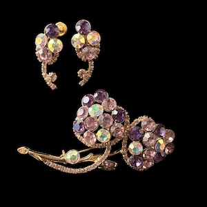 Puede incluir: Un conjunto de joyería compuesto por un broche y un par de pendientes. Las piezas presentan monturas de metal en tono dorado adornadas con grupos de cristales redondos y facetados en colores púrpura, rosa e irisado con formas florales.
