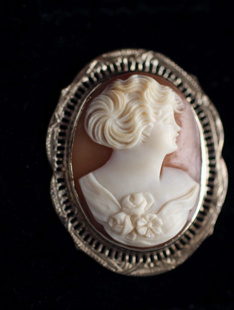 May include: A cameo brooch featuring a carved portrait of a woman with a rose in her hair, set in a silver tone metal frame.
