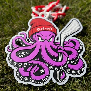 Op de afbeelding: Een paarse octopus met een rode hoed met de tekst "Detroit" erop, die een witte hockeystick vasthoudt. De octopus heeft zwarte ogen en een dreigende uitdrukking.