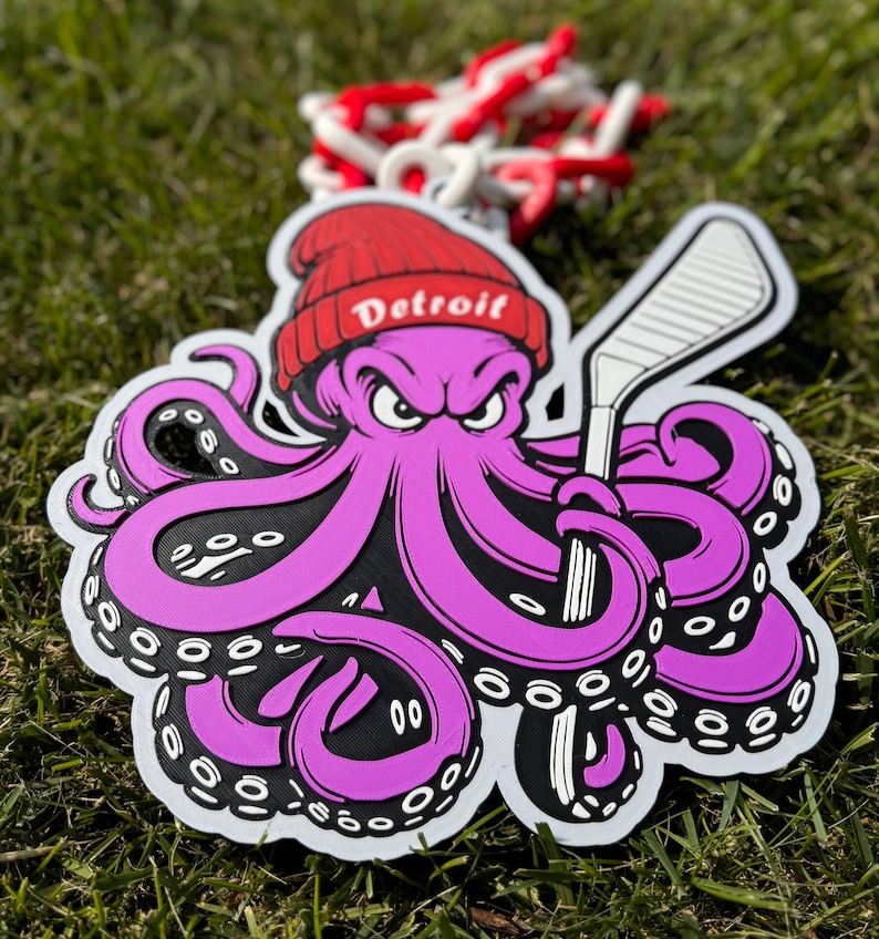 Op de afbeelding: Een paarse octopus met een rode hoed met de tekst "Detroit" erop, die een witte hockeystick vasthoudt. De octopus heeft zwarte ogen en een dreigende uitdrukking.