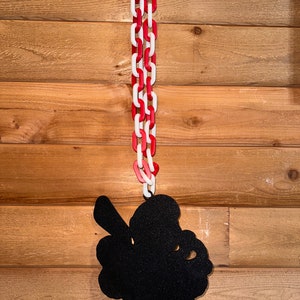 Op de afbeelding: Een zwart silhouet van een klavertjevier dat hangt aan een rood-witte ketting op een houten paneelwand.