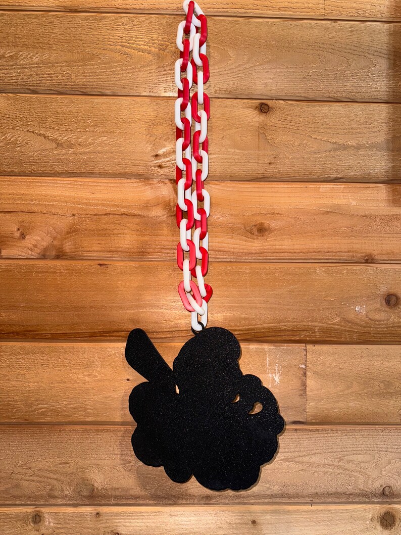 Op de afbeelding: Een zwart silhouet van een klavertjevier dat hangt aan een rood-witte ketting op een houten paneelwand.