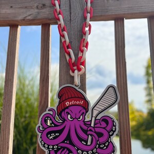 Op de afbeelding: Een paarse octopus met een rode hoed met het woord "Detroit" erop, houdt een hockeystick vast. De octopus hangt aan een rood-witte ketting.