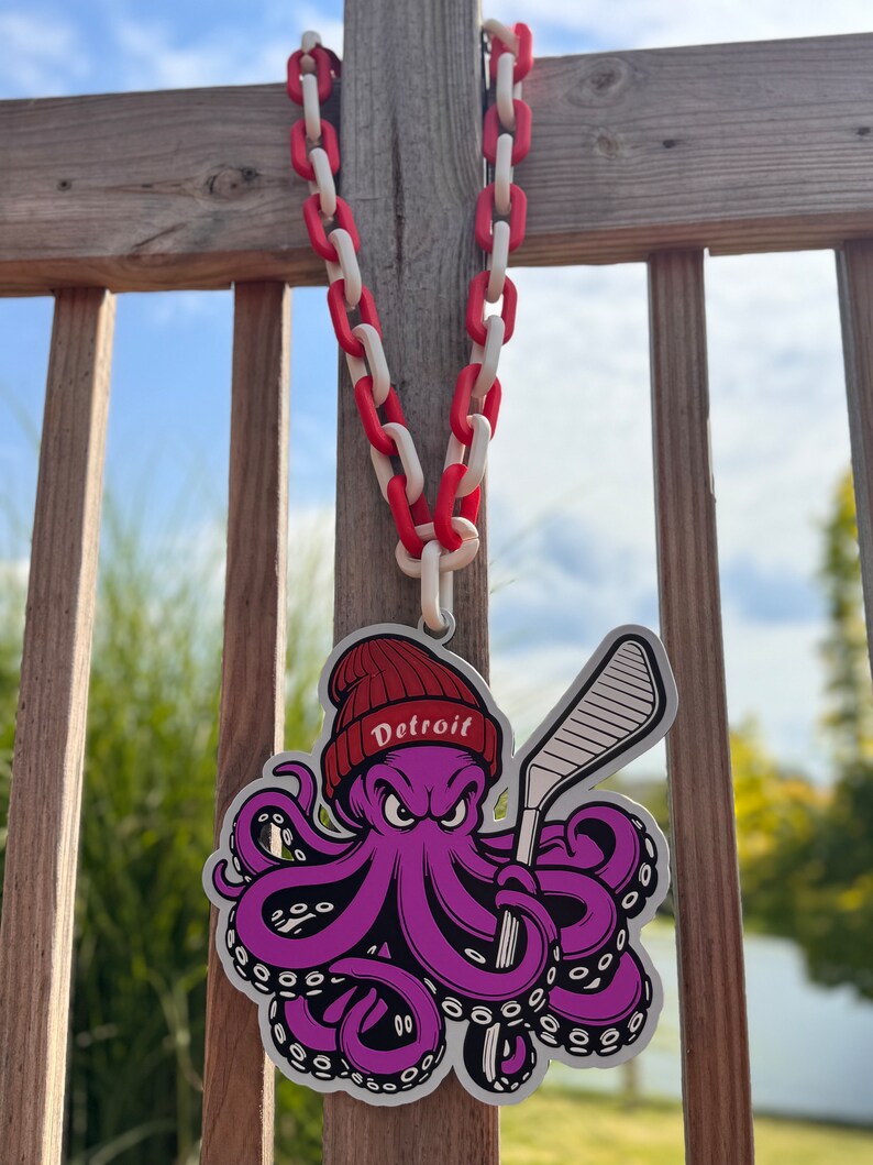Op de afbeelding: Een paarse octopus met een rode hoed met het woord "Detroit" erop, houdt een hockeystick vast. De octopus hangt aan een rood-witte ketting.