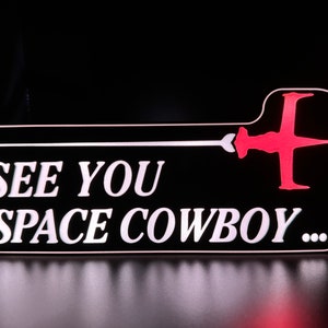 May include: A black and white sign with the text "See You Space Cowboy..." in white lettering. A red spaceship silhouette is in the top right corner.