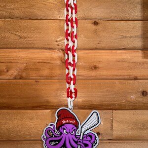 Op de afbeelding: Een paarse octopus die een rode hoed draagt met het woord "Detroit" erop, en een hockeystick vasthoudt. De octopus hangt aan een rood-witte ketting.