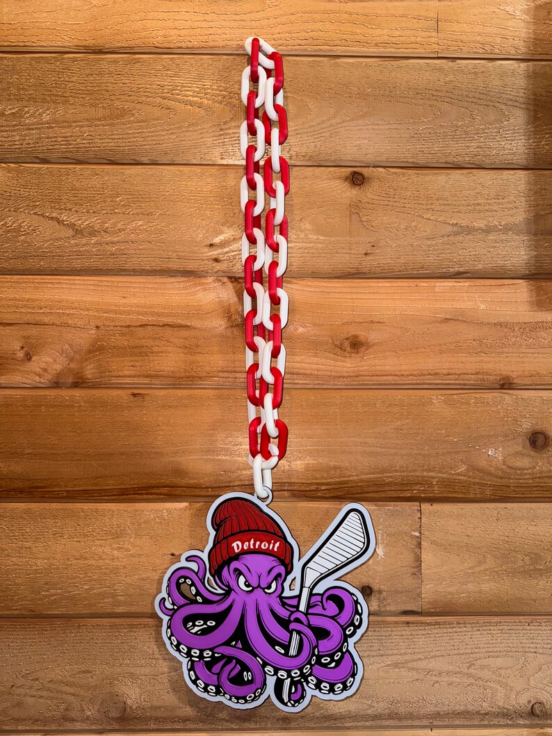 Op de afbeelding: Een paarse octopus die een rode hoed draagt met het woord "Detroit" erop, en een hockeystick vasthoudt. De octopus hangt aan een rood-witte ketting.