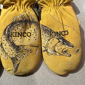 May include: A pair of yellow Kinco mittens featuring black ink illustrations of trout fish and the text KINCO printed on the front. The mittens have black knit cuffs and a small black tag labeled Heatkeep on the side of the left mitten.