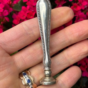 May include: A silver antique wax seal stamp with a decorative handle and a round base. The seal is held in a hand, with a ring visible on the finger. The background features vibrant red flowers.