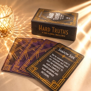 May include: A black and gold oracle deck box titled Hard Truths A Fate Altering Oracle Deck with 66 cards. Several cards are fanned out, featuring a purple and gold geometric border. One card displays the text Shadow Side and advice in white.