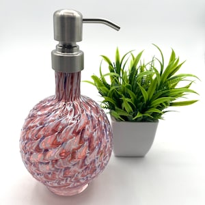 Puede incluir: Un dispensador de jabón de vidrio con un diseño de espiral rosa y blanco con una bomba de acero inoxidable. El dispensador está sobre una superficie blanca junto a una pequeña planta en maceta.