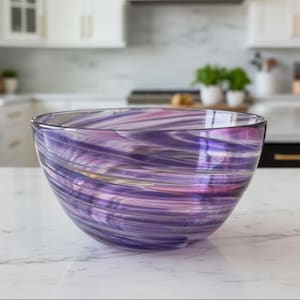May include: A large, clear glass bowl with a swirling pattern of purple, pink, and white. The bowl is set on a white countertop in a kitchen setting. The bowl's design is visible through the glass.