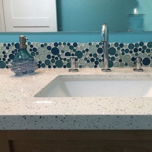 Puede incluir: Un tocador de ba&ntilde;o blanco con un lavabo de porcelana blanca y tres grifos cromados. La encimera tiene un acabado moteado. Un salpicadero de azulejos de mosaico azul y verde est&aacute; detr&aacute;s del lavabo.
