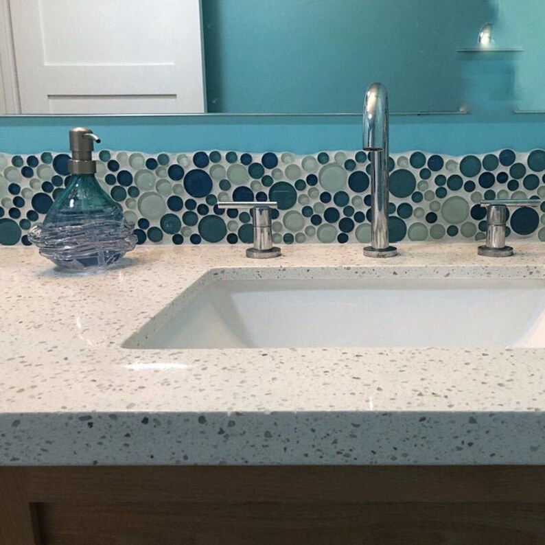 Puede incluir: Un tocador de ba&ntilde;o blanco con un lavabo de porcelana blanca y tres grifos cromados. La encimera tiene un acabado moteado. Un salpicadero de azulejos de mosaico azul y verde est&aacute; detr&aacute;s del lavabo.