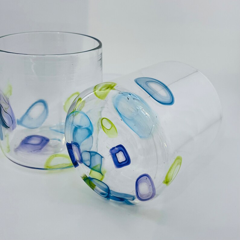 May include: Two clear glass tumblers with colourful abstract designs. One tumbler is upright, the other is on its side. The designs include blue, green, and purple shapes. The tumblers are on a white surface.