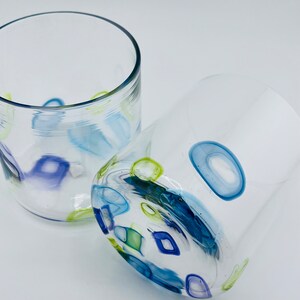 May include: Two clear glass tumblers with colourful abstract designs. The designs include blue, green, and purple shapes. The tumblers are on a white background. The glass has a rounded shape.